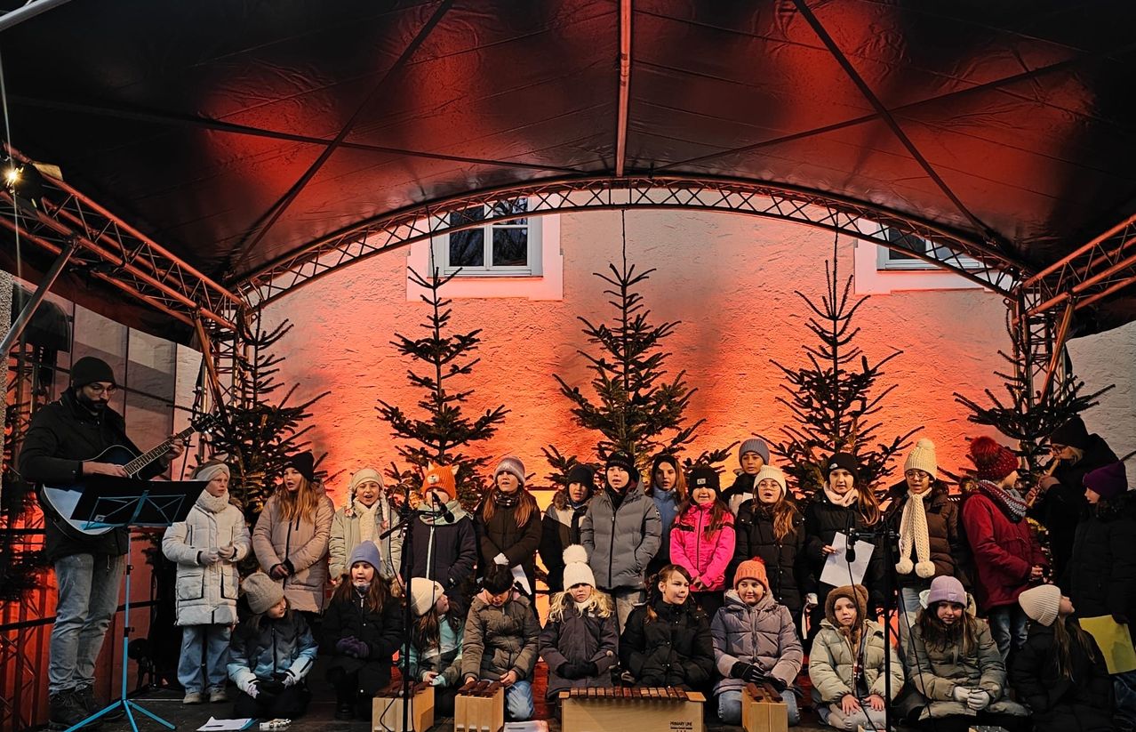 Ein Kinderchor tritt vor einem Hintergrund aus Weihnachtsbäumen und einer leuchtend orangen Wand auf, mit verschiedenen Musikinstrumenten im Vordergrund.