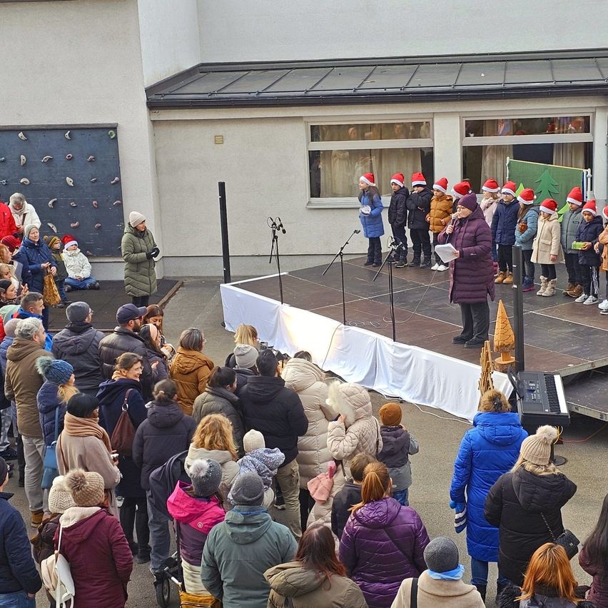 Eine Gruppe von Menschen versammelt sich vor einem Gebäude, einige tragen Mützen, während einige Kinder auf der Bühne mit einer Frau, die ein Mikrofon hält, singen.