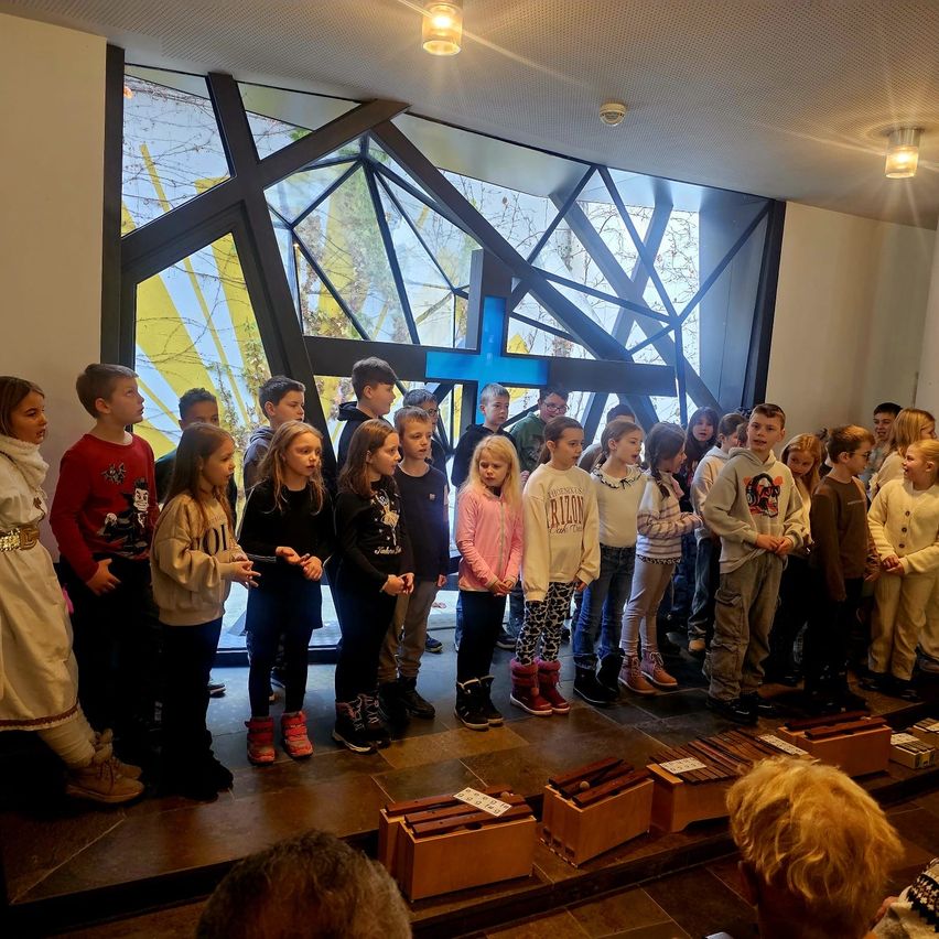 Eine Gruppe von Kindern steht in einer Kirche mit einem großen Kreuz hinter ihnen. Einige singen, während andere zusehen. Der Raum hat einen gefliesten Boden und geometrische Glasfenster.