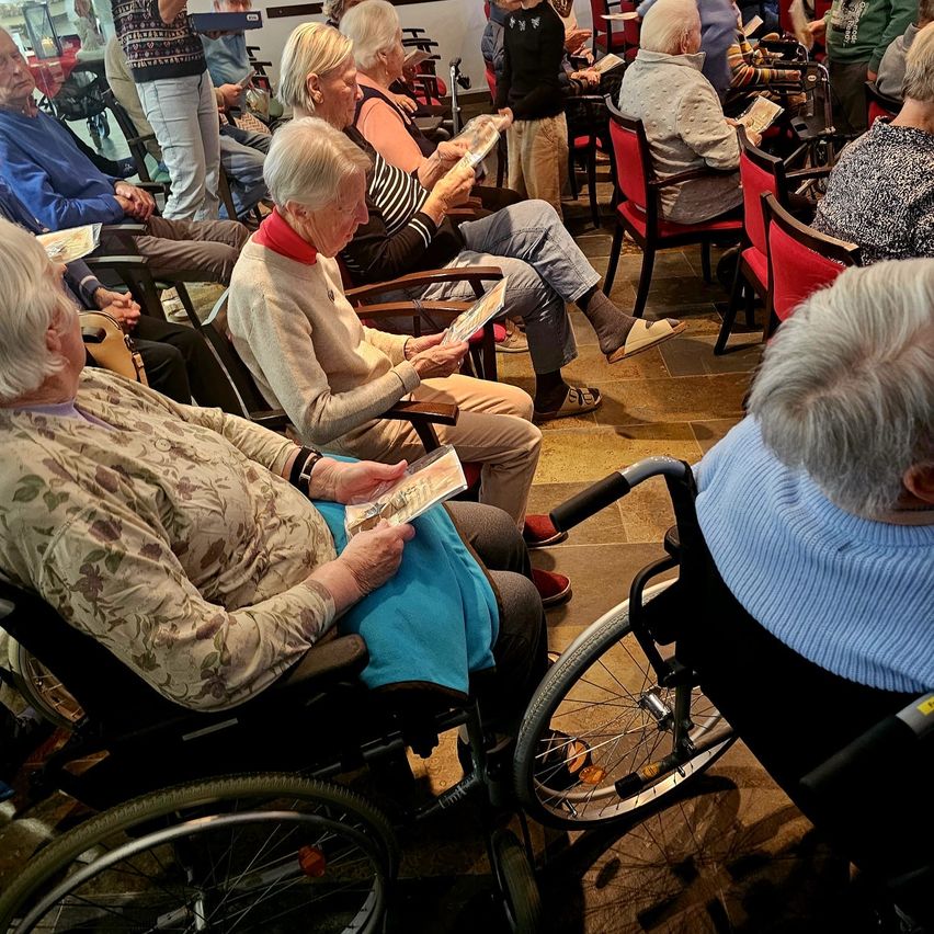 Mehrere ältere Menschen in Rollstühlen und Stühlen sitzen in einem Raum und lesen Bücher und Papiere. Der Raum hat einen gefliesten Boden und rote Stühle.