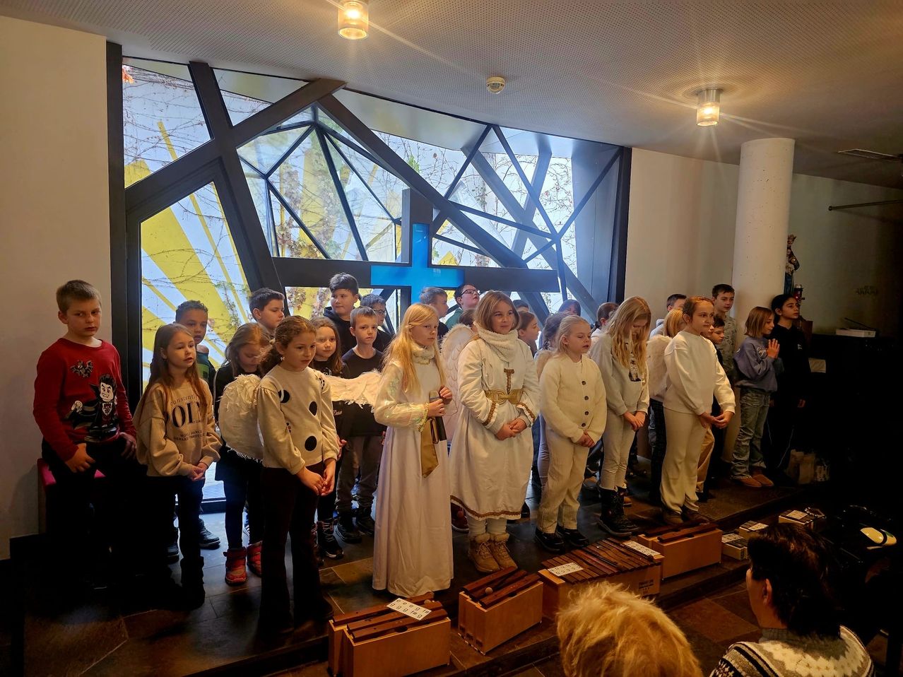 Eine Gruppe von Kindern versammelt sich in einer Kirche, gekleidet in Weiß und einige in Engelskostümen, halten Musikinstrumente. Sie stehen vor einem großen Fenster mit einem blauen Kreuz.