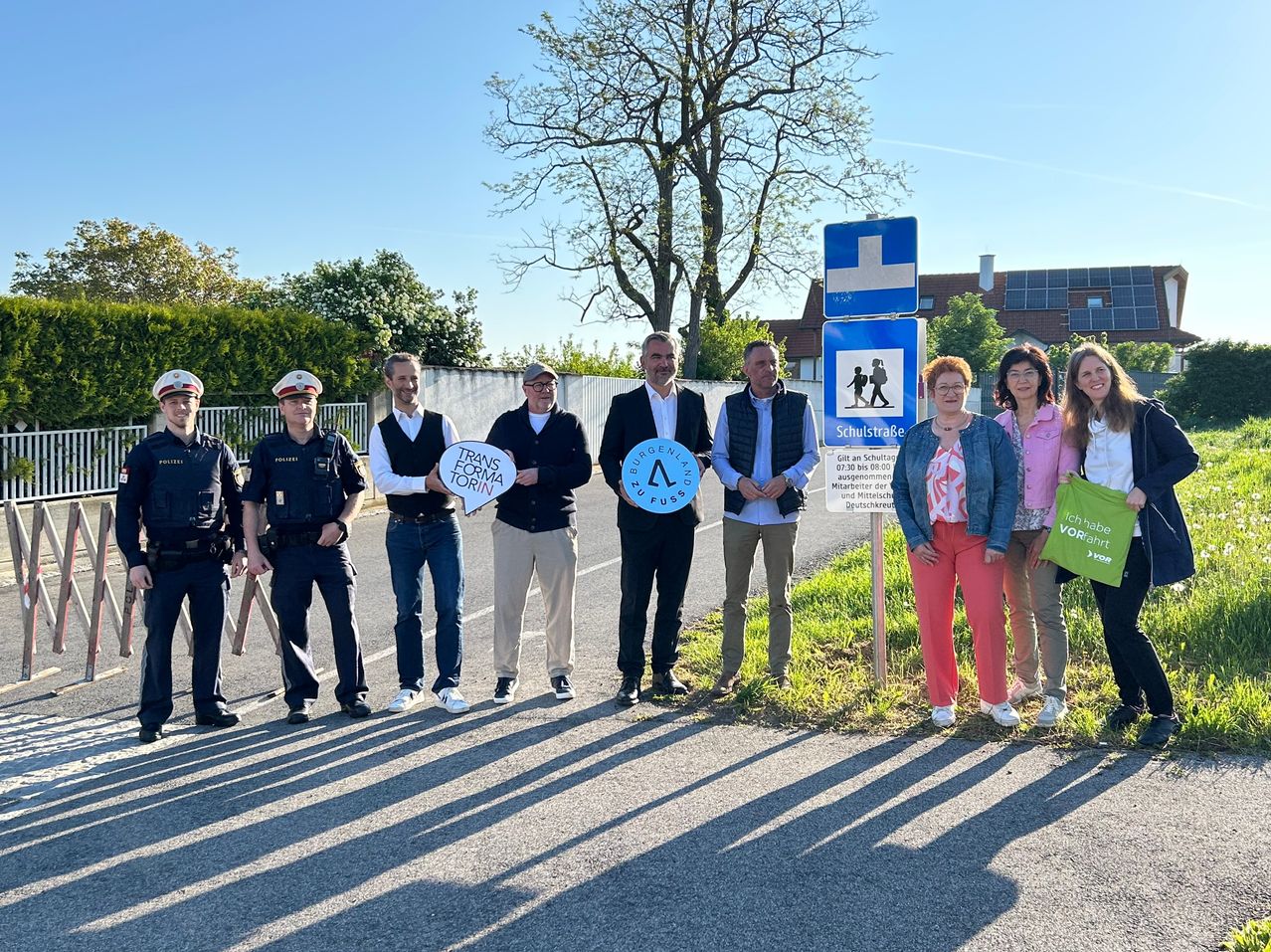 Mehrere Personen stehen am Straßenrand, mit einem blauen Schild, das "Schulstraße" und ein weißes Schild mit einem Kreuz sagt. Einige von ihnen halten ein Schild, das "TRANS FORMA TORiN" sagt.