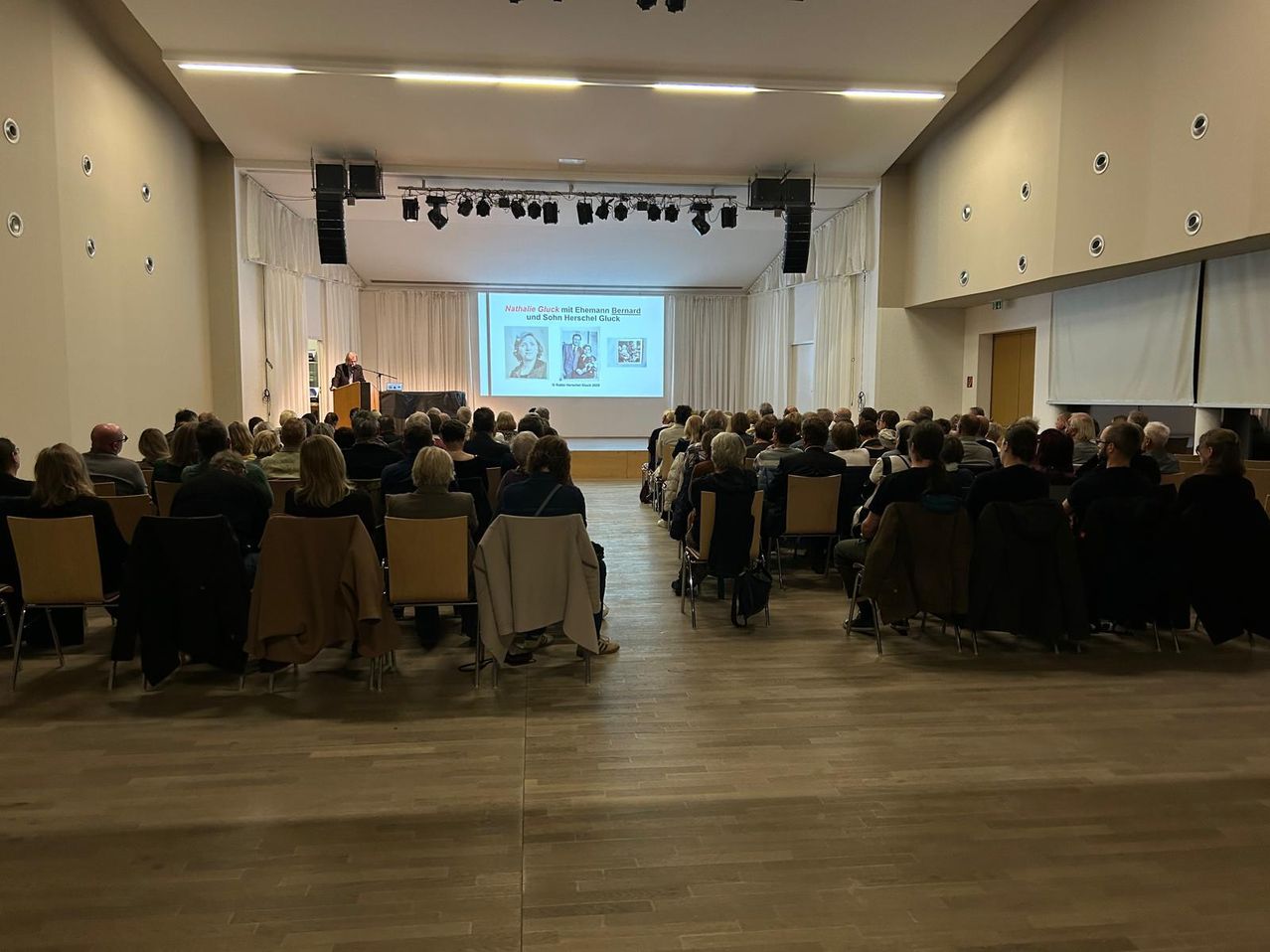 Ein Saal mit einer Projektionsleinwand, auf der eine Präsentation mit Bildern von Personen gezeigt wird. Ein Mann spricht an einem Rednerpult. Das Publikum sitzt auf Stühlen.
