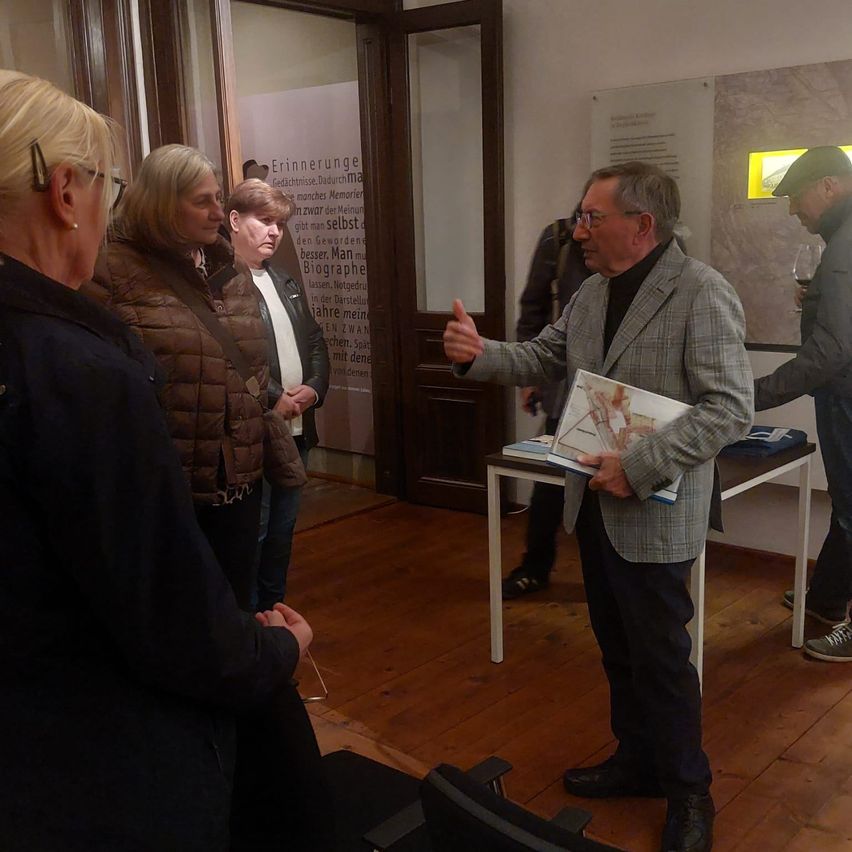 Ein Mann in einem Anzug hält eine Präsentation für eine Gruppe von Frauen, eine hält ein Glas Wein, in einem Raum mit Holzböden.