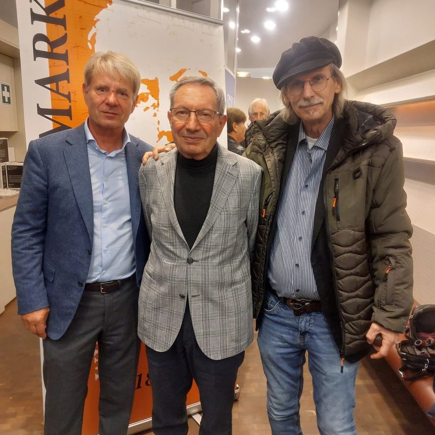 Drei Männer posieren für ein Foto vor einem Banner im Hintergrund. Der Mann auf der linken Seite trägt einen blauen Anzug, der in der Mitte einen grauen Blazer und der rechte eine grüne Jacke.