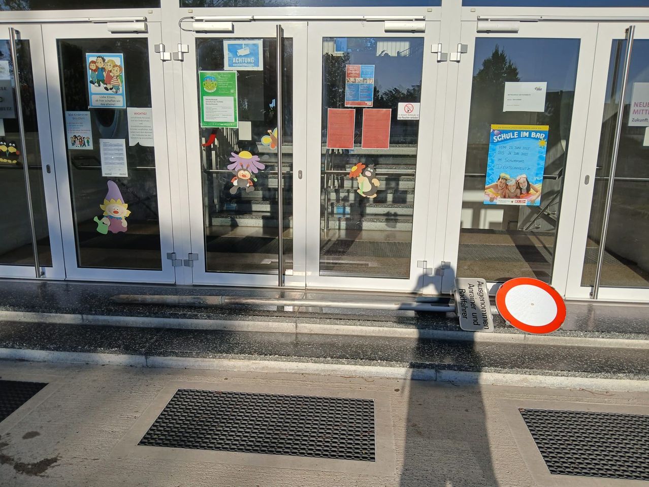 Ein Gebäudeeingang mit Glastüren hat mehrere Hinweise und Schilder darauf. Es gibt zwei Mattebereiche auf dem Bürgersteig und ein rotes und weißes Schild.