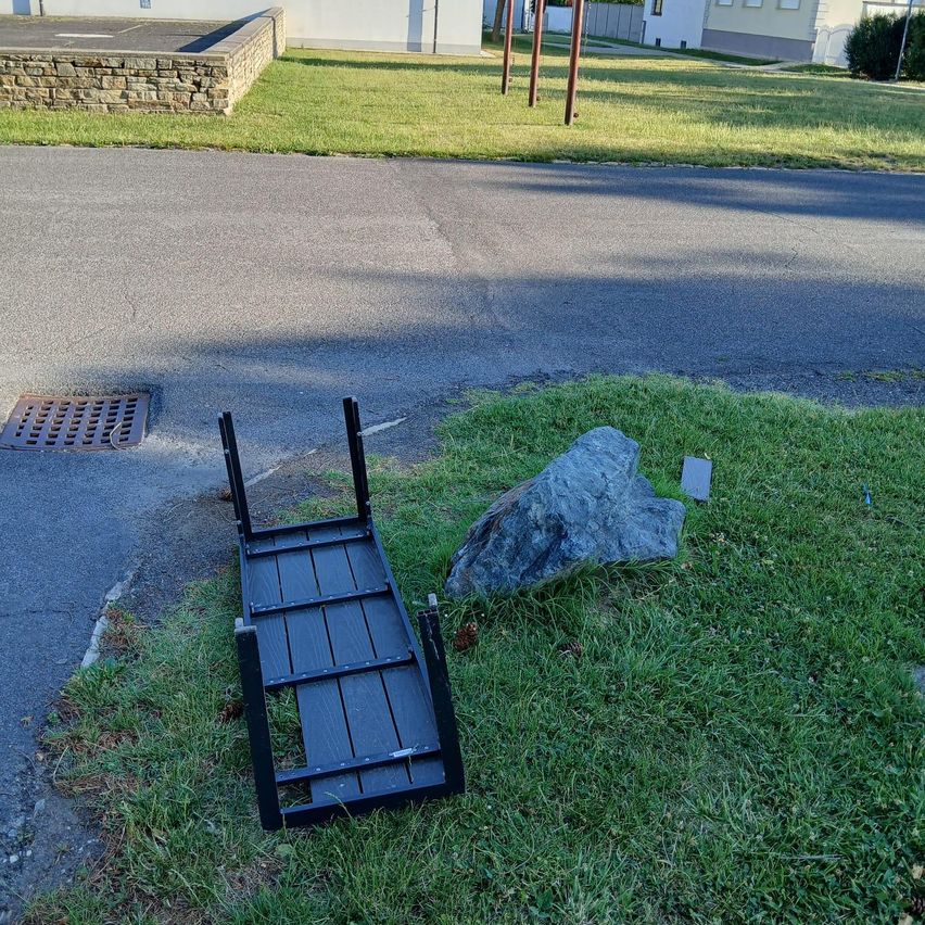 Eine schwarze faltbare Bank liegt auf dem Rasen in der Nähe einer Straße mit einer Steinmauer im Hintergrund.
