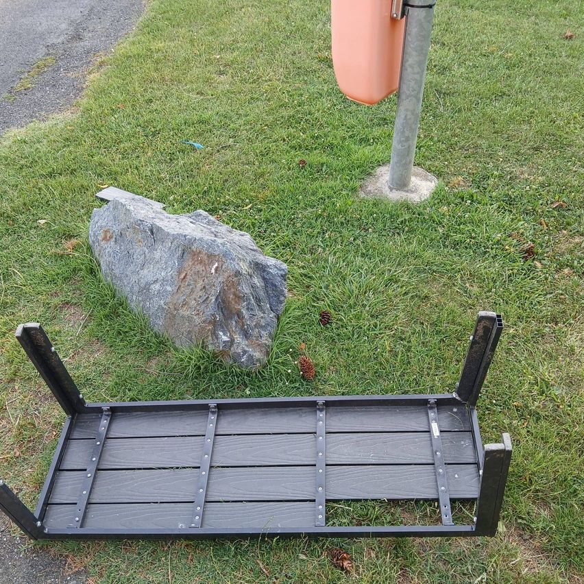 Eine schwarze Metallbank liegt neben einem großen Felsen auf dem Gras in der Nähe eines Pfostens mit einem Mülleimer.