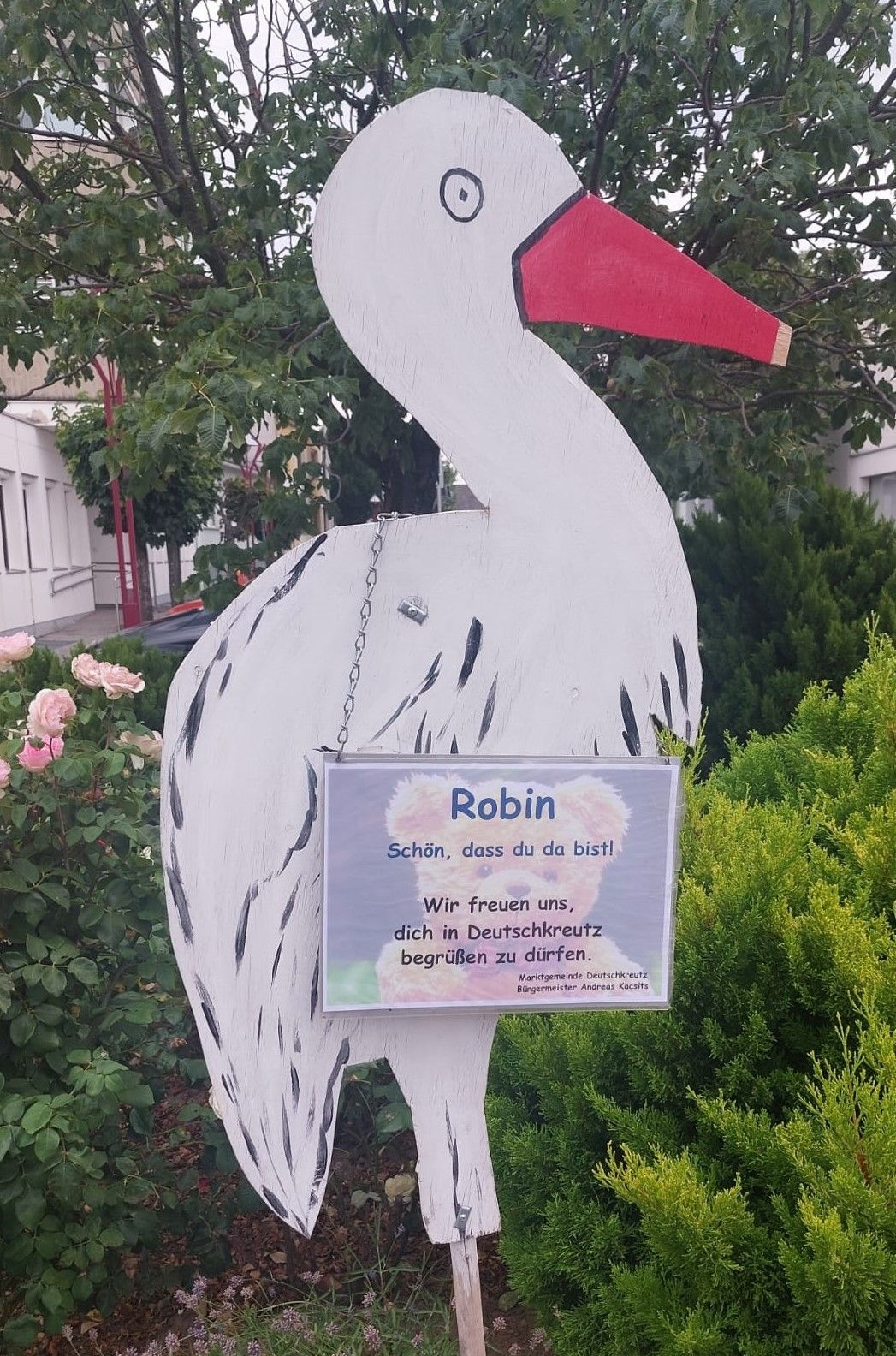 Eine Storchskulptur mit einem Schild, auf dem 'Robin' steht. Das Schild enthält eine Nachricht auf Deutsch. Dahinter befinden sich Büsche, rosa Blumen und ein Gebäude mit Glasfenstern.