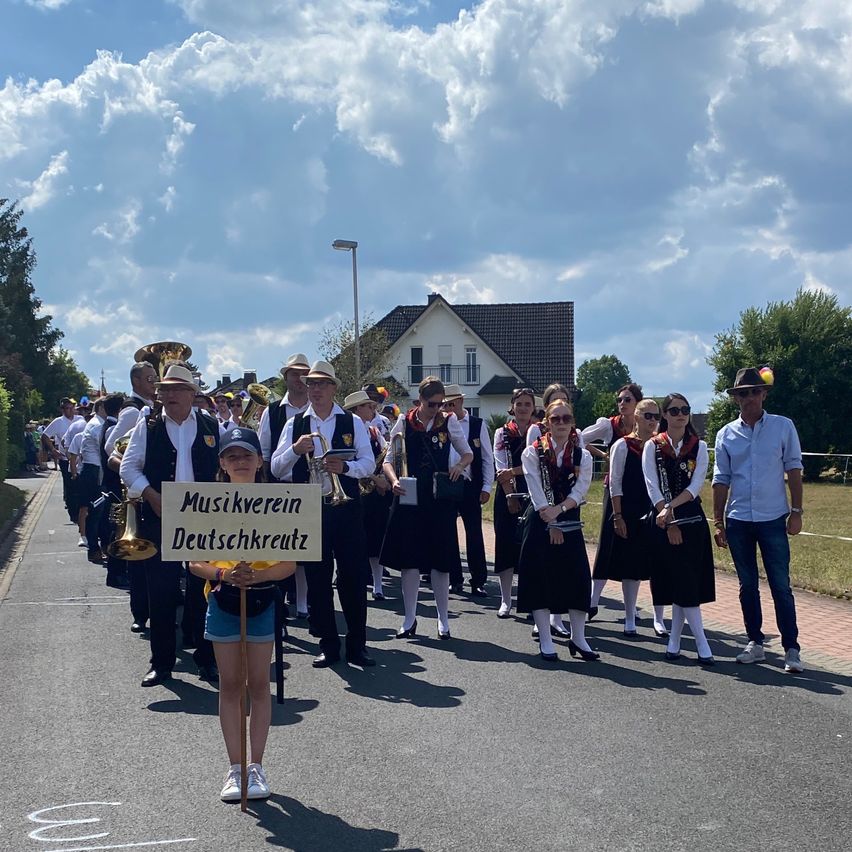 Eine Gruppe von Menschen, möglicherweise eine Blaskapelle, geht auf der Straße. Ein Mädchen hält ein Schild mit den Worten 'Musikverein Deutschkreutz'. Dahinter halten Menschen Musikinstrumente. Ein Mann in einem blauen Hemd steht auf der rechten Seite. Es gibt Häuser am Straßenrand.