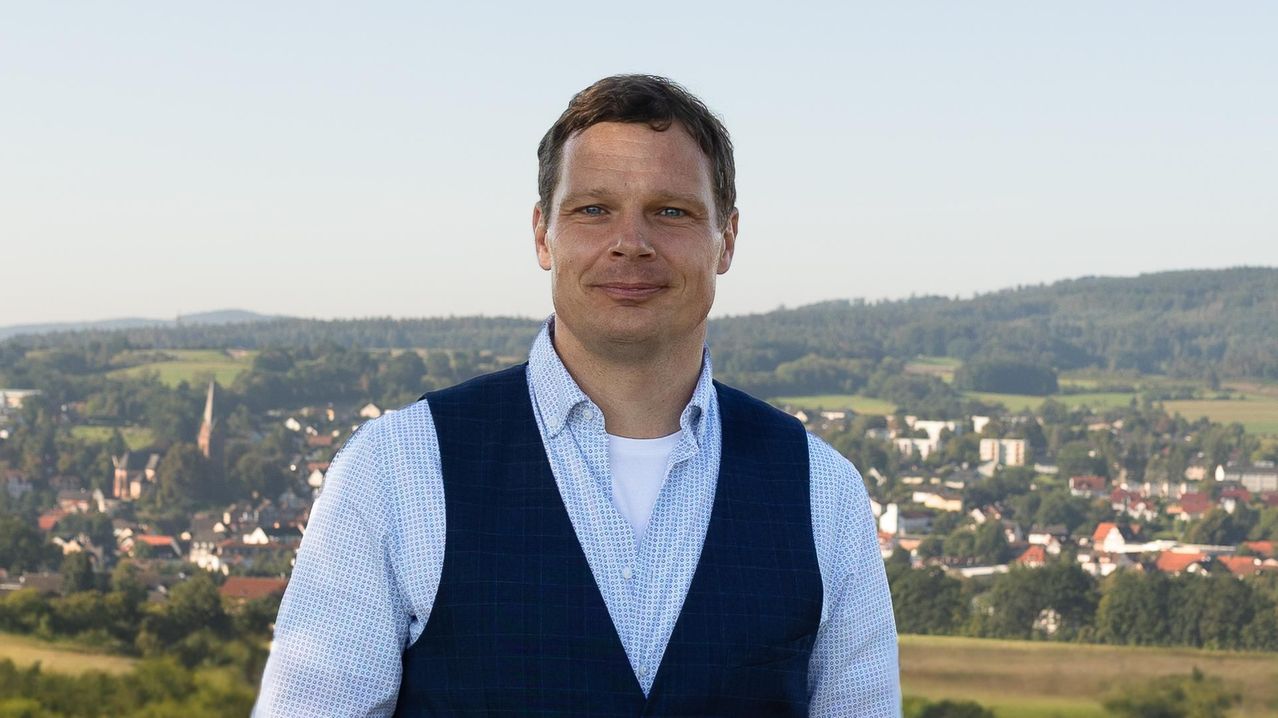 Ein Mann steht im Freien, lächelt und hat eine Stadt und Hügel im Hintergrund. Er trägt eine blaue Weste über einem weißen Hemd.