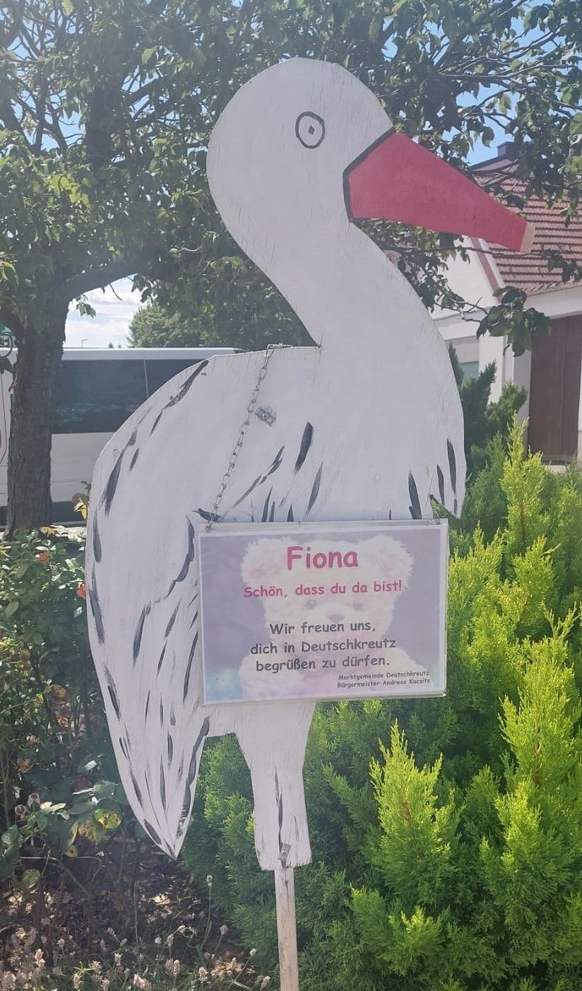Eine Storchstatue mit einem Schild mit dem Namen 'Fiona' in rosa Buchstaben begrüßt Besucher in einem Garten. Dahinter sind ein Haus mit rotem Dach und ein weißer Bus zu sehen.