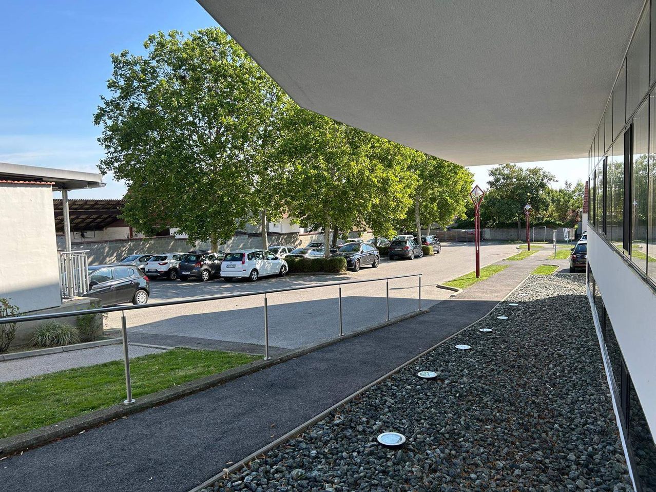 Ein Parkplatz mit mehreren Autos unter Bäumen. Das Gelände ist von einer Umzäunung und Straßenlaternen umgeben.