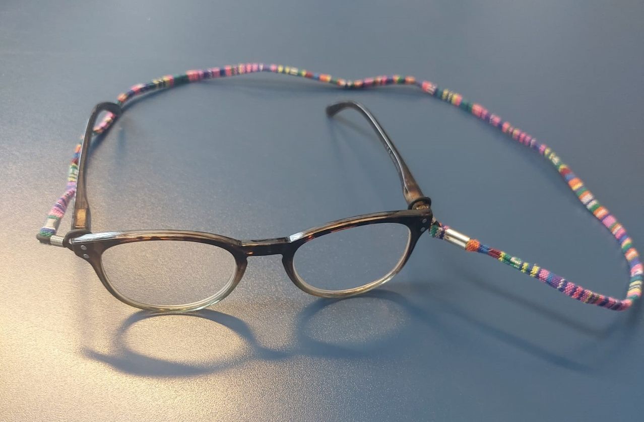 Ein Paar schwarzer Brille mit einer bunten Perlenschnur liegt auf einer grauen Oberfläche. Die Schnur hat verschiedene Farben und ist mit der Brille verbunden.