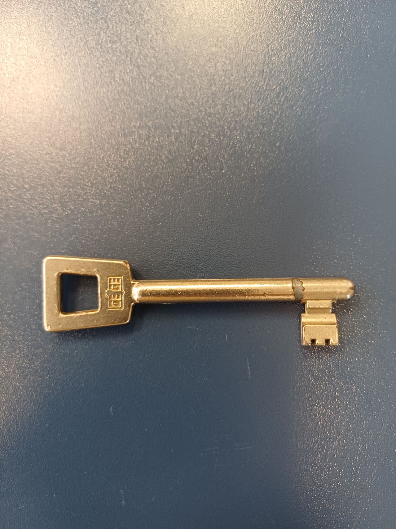 Ein goldener Schlüssel mit den eingeprägten Buchstaben DE GE liegt auf einer grauen Oberfläche.