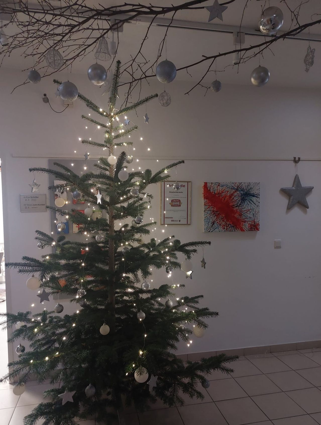 Ein geschmückter Weihnachtsbaum mit Lichtern und Ornamenten steht in einem Raum mit weißen Wänden, Fliesenboden und einer an der Wand montierten Malerei.