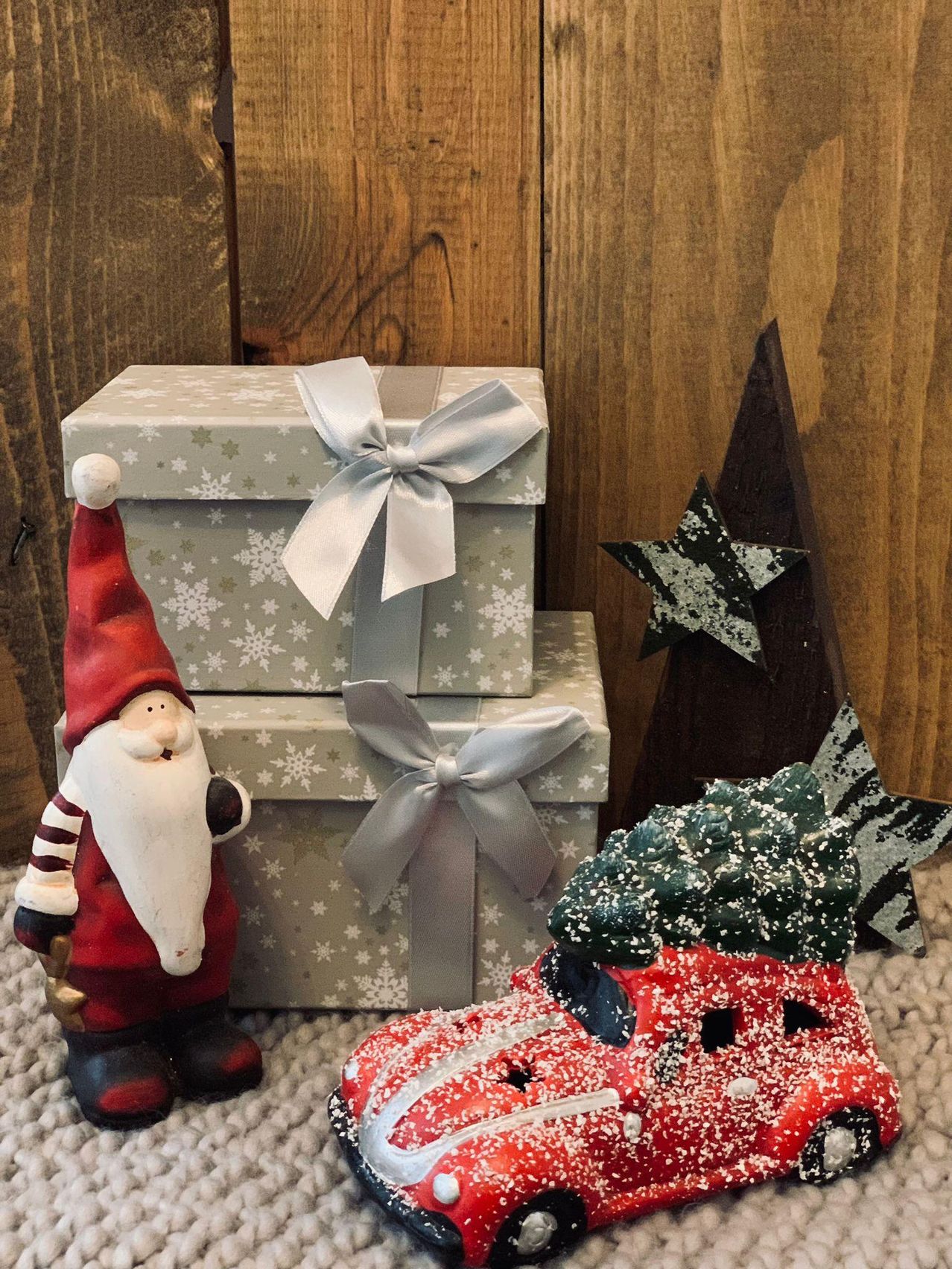Eine kleine Weihnachtsmann-Figur steht neben einem Stapel von drei verpackten Geschenken, mit einem roten Auto und einer sternförmigen Dekoration in der Nähe. Der Hintergrund ist eine Holzwand.