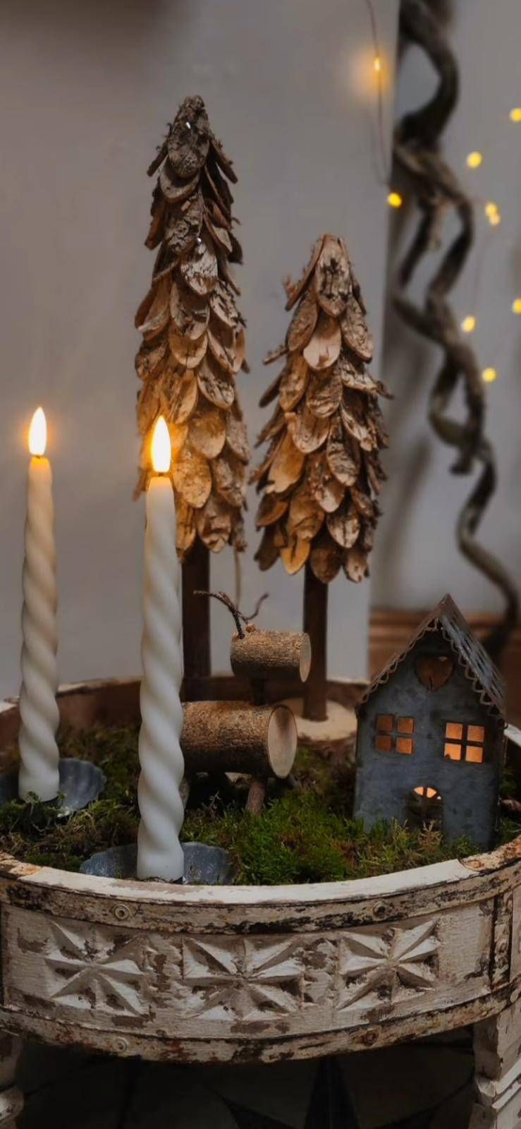 Eine dekorative Szene mit zwei brennenden weißen Kerzen, dekorativen Holz-Kiefernbaum-Figuren, einem Baumstamm und einem kleinen beleuchteten Haus aus Metall.