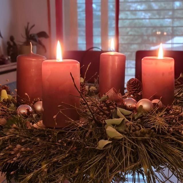 Ein Kranz aus Tannenzapfen und Kiefernnadeln umgibt vier brennende, rosa Kerzen und vier silberne und goldene Weihnachtskugeln. Eine Pflanze und Vorhänge sind im Hintergrund.