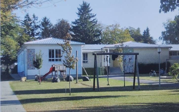 Ein weißes Haus mit Metalldach hat einen roten Rutschen, einen Schaukelplatz und ein Fahrrad in der Nähe. Bäume und Büsche im Hintergrund.