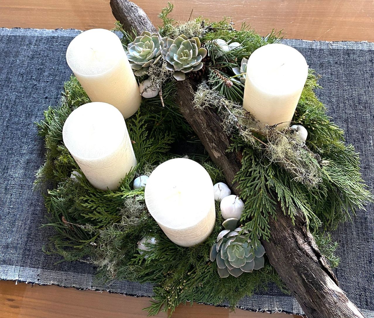 Ein rustikaler Kranz aus einem Baumzweig, geschmückt mit grünem Laub, Sukkulenten und vier weißen Kerzen.