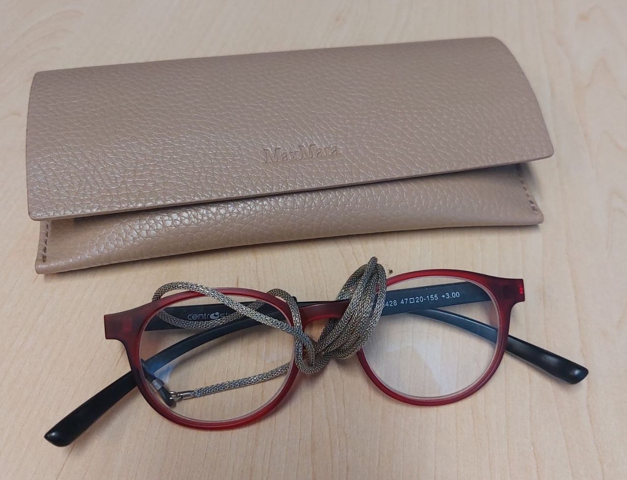 A pair of red-framed glasses with a black frame lies on a wooden surface, next to a beige leather case.