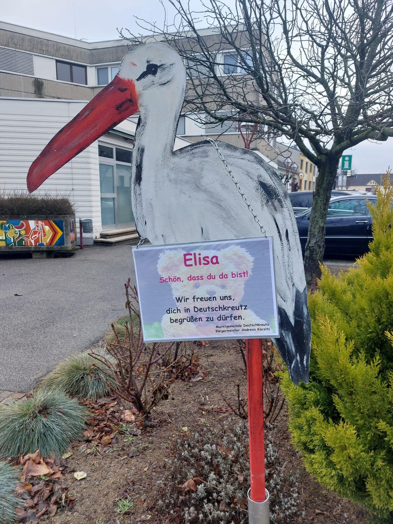 Eine Storchstatue namens Elisa steht in einem Garten mit einem Schild, das Besucher begrüßt, sie in Deutsch zu streicheln. Dahinter steht ein Gebäude mit einem Wandbild und einem Baum.