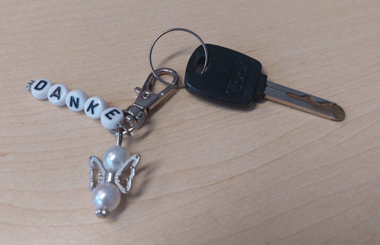 Ein Schlüsselanhänger mit den Buchstaben N, K und E, befestigt an einem Schlüssel mit einem Schmetterlingsanhänger und Perlen auf einer Holzoberfläche.