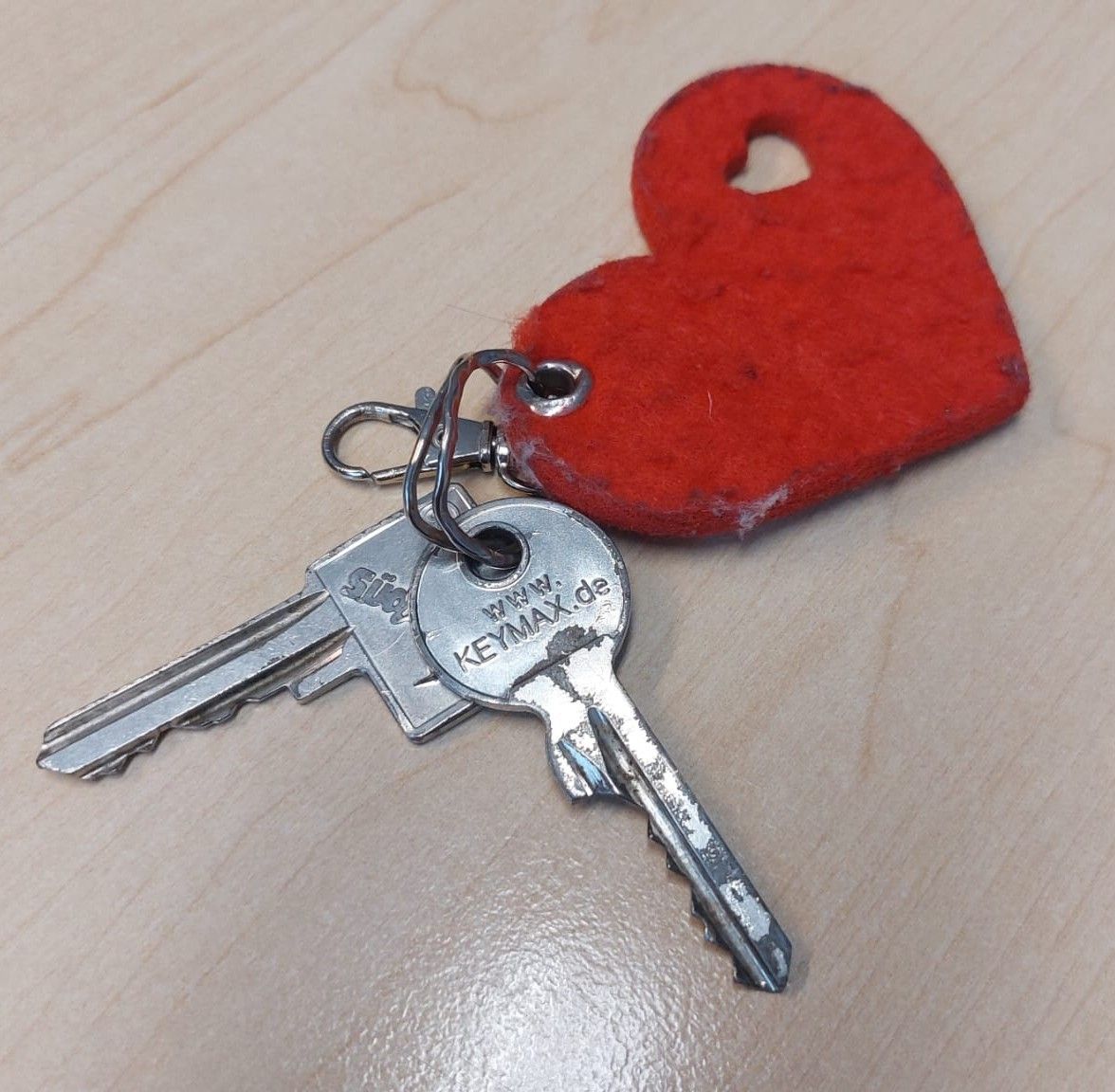 Two silver keys attached to a red heart-shaped keyring are placed on a wooden surface. The keys have 'WWW' and 'KEYMAX.de' engraved.