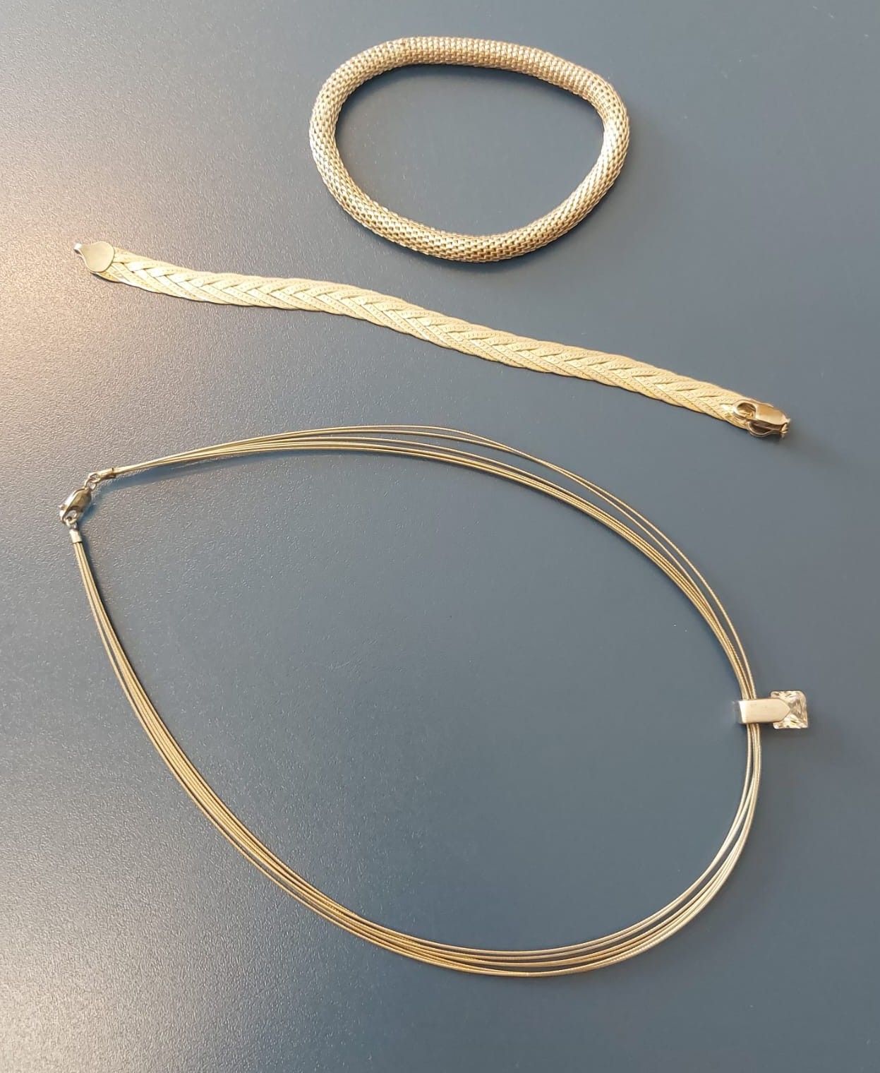 Three gold necklaces on a gray surface. The top one is a coiled necklace. The middle one is a braided necklace. The bottom one has a square pendant.