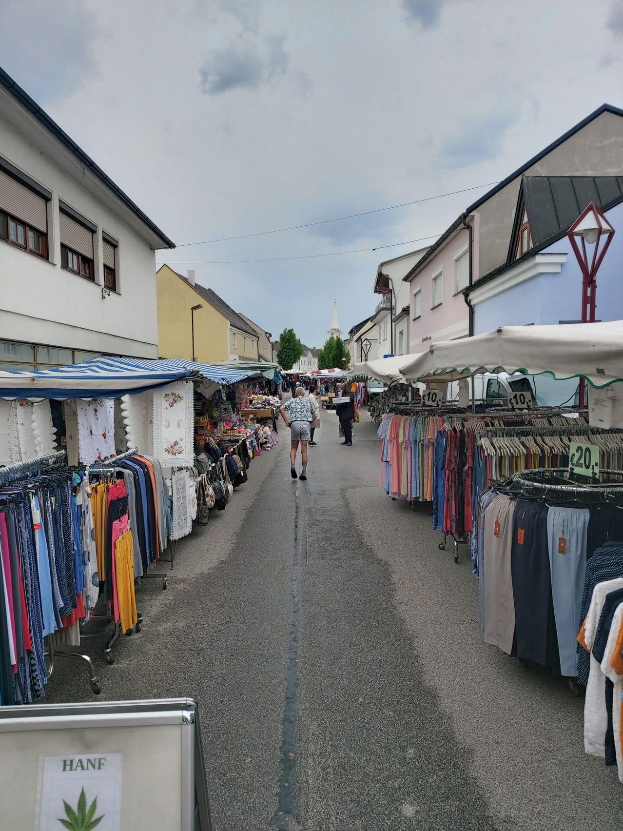An outdoor market street with shops on both sides. Clothing items are displayed on the side of the road. A person walks on the street. Trees and buildings are visible in the background.