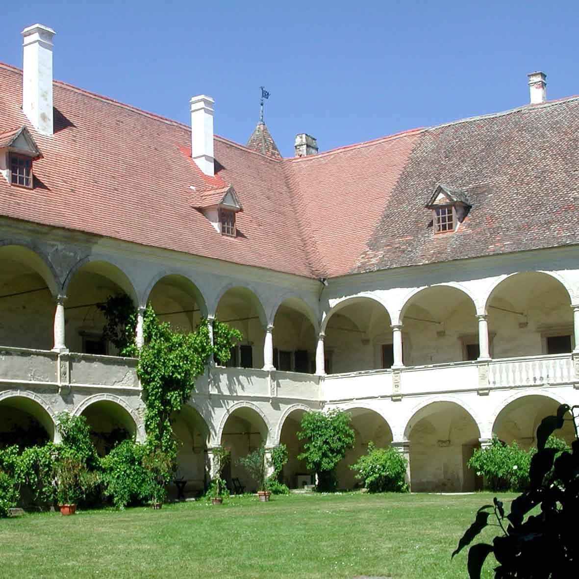 A courtyard of an old building with arched windows and a red tiled roof. Green grass and plants are in front.
