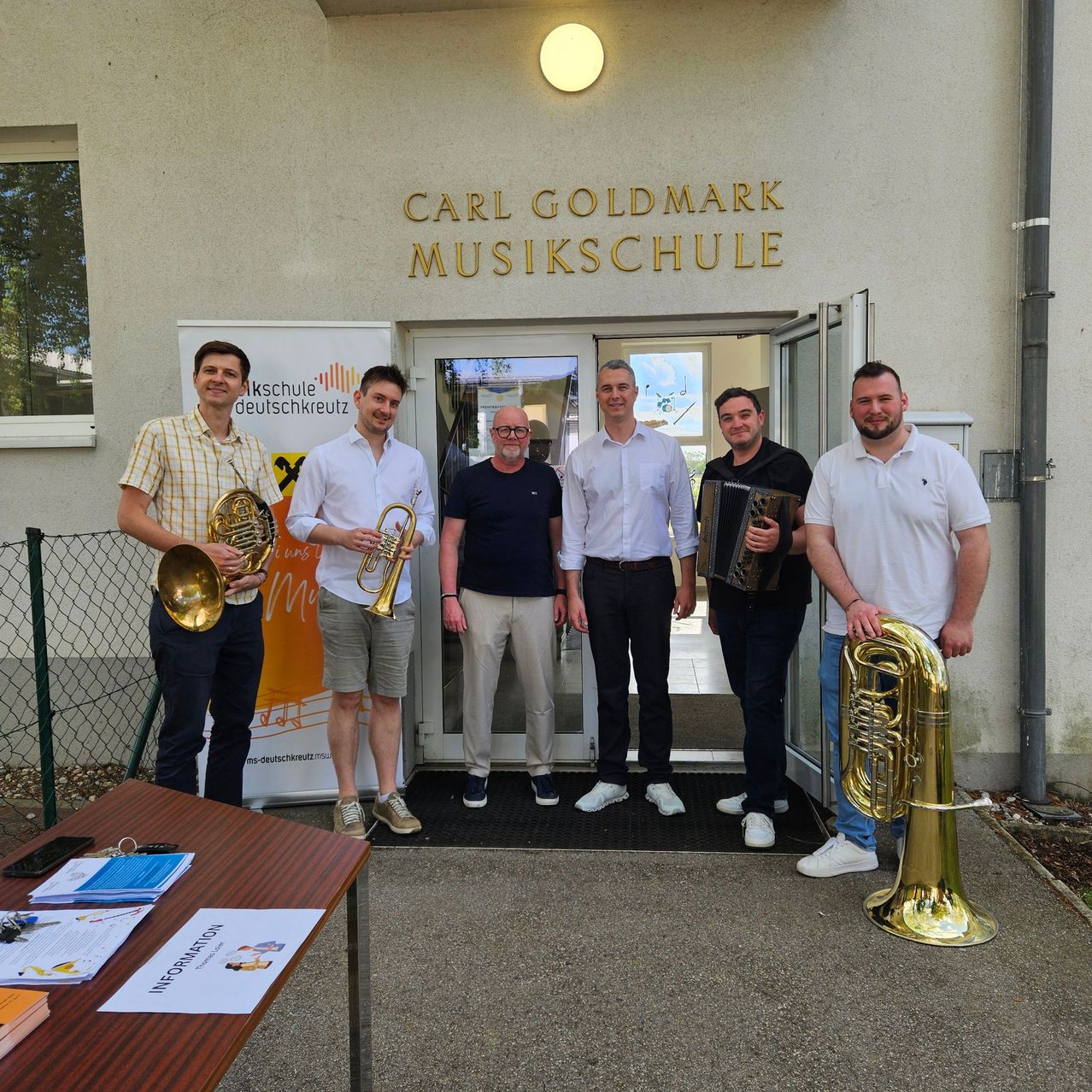 Five men stand in front of a music school, each holding a brass instrument. The building's sign reads Carl Goldmark Musikschule. Papers and a phone are on a table in front of them.