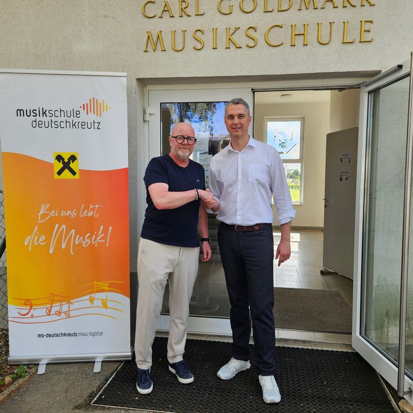 Two men stand in front of a music school entrance, smiling and posing for a photo. A banner next to them reads 'Beinus lebt die Musik!' and features musical notes.