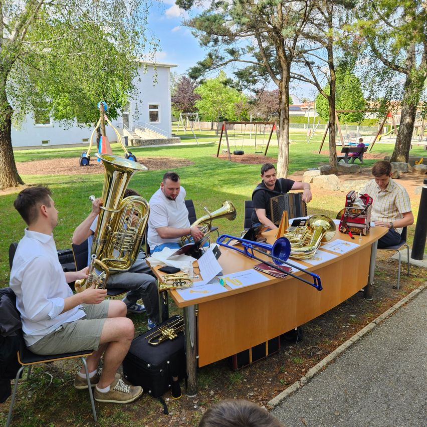 Five men are seated around a table, playing various brass instruments in a park setting. Papers and sheet music are on the table. A building and playground equipment are visible in the background.