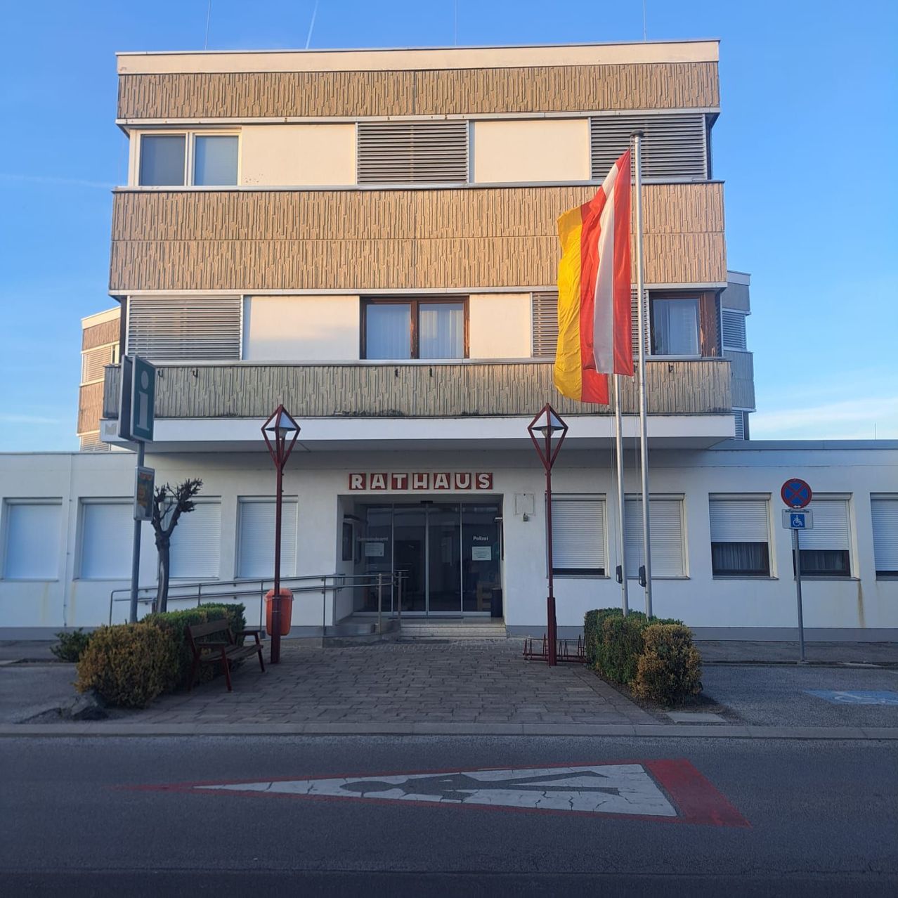 A white building with several floors and a red sign reading 'Rathaus'. A flag is hoisted on a pole. Lamps are in front of the entrance. A bench is placed on the pathway.