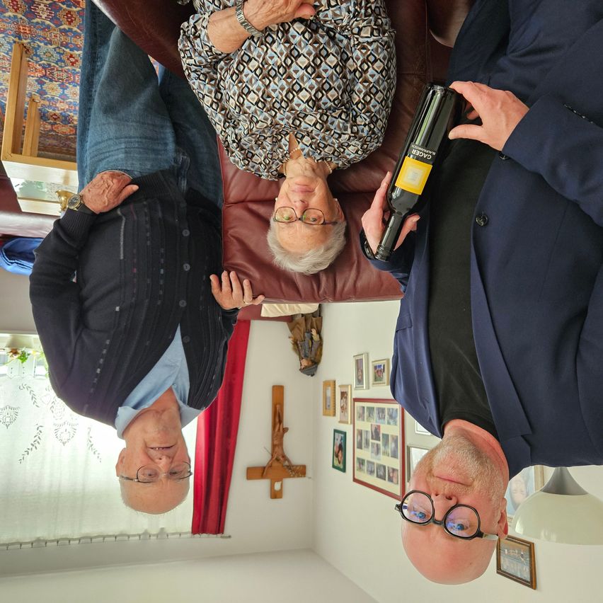 Three people are upside down, holding a bottle of wine. The woman wears glasses and a patterned top. The man on the left wears a watch and a sweater. The man on the right wears a blue coat and glasses. They are in a room with picture frames and a cross on the wall.
