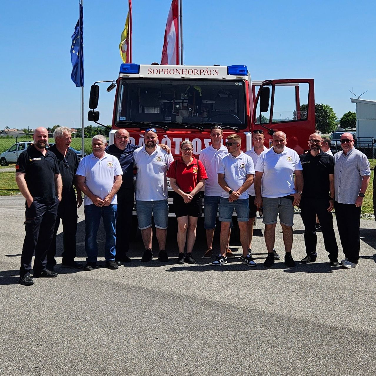 Eine Gruppe von Männern steht vor einem roten Feuerwehrauto mit der Aufschrift 'Sopronhorpacs'. Sie tragen weiße Hemden und Shorts. Eine Frau steht in der Mitte, die ein rotes Shirt trägt. Hinter ihnen stehen Fahnen, und in der Ferne sind Bäume und Autos zu sehen.