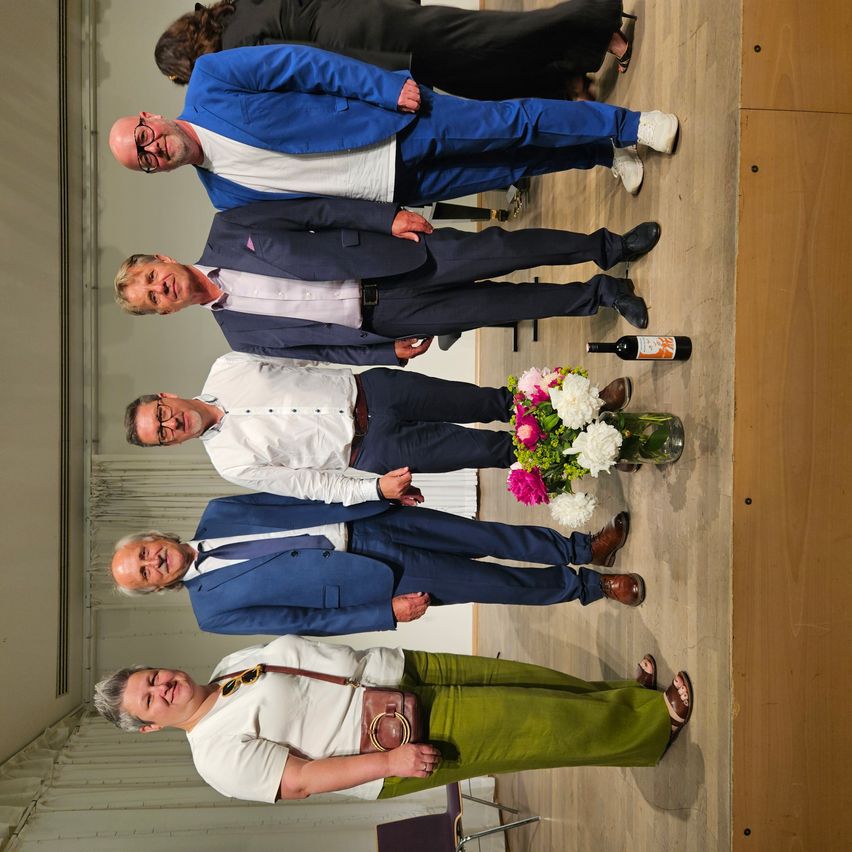 Five men are posing for a photo, all dressed formally, standing on a wooden floor. A bouquet of flowers and a bottle of wine are nearby.