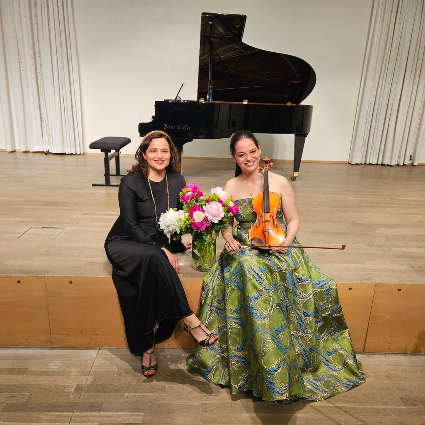 Two women are sitting on a stage with a piano and flowers behind them. One holds a violin.