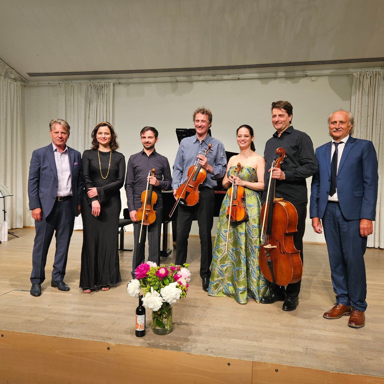 A group of musicians stands on stage, each holding a violin or cello, smiling for a photograph. A piano is behind them, with a bouquet of flowers and a wine bottle in the foreground.