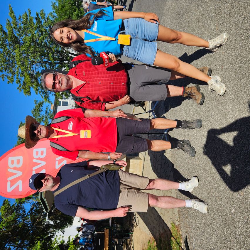 Four people are posing for a photo on a street, with a red banner reading BUS behind them. They are wearing casual clothing and have smiles on their faces.