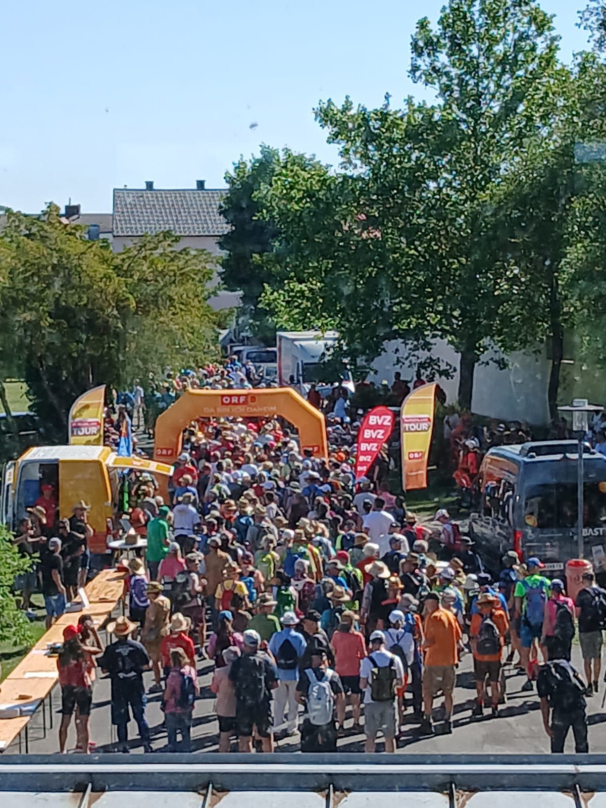 A large crowd of people, some wearing hats and backpacks, are gathered in a street, possibly for an event. The area is lined with trees and street lights. Banners are visible in the background.