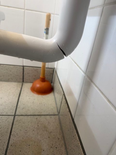 A white pipe with a crack is attached to a tiled wall, with a plunger placed underneath it.