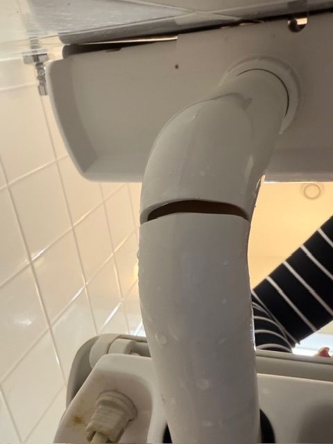 A white toilet part with a broken section and a plug is mounted on a tiled wall. The background includes a striped shirt and a bathroom light.