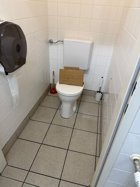 A bathroom with tiled floor and white walls. A toilet is in the corner with a cardboard box on its seat. A toilet paper dispenser and a plunger are nearby.