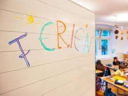 A classroom with children seated at desks, a white wall has colorful words and drawings, including a sun, and windows with views of the outdoors.