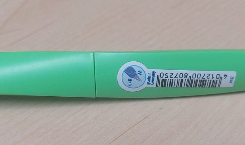 A close-up of a green pen with a white label displaying the brand 'L+R', made in Germany, and a barcode.