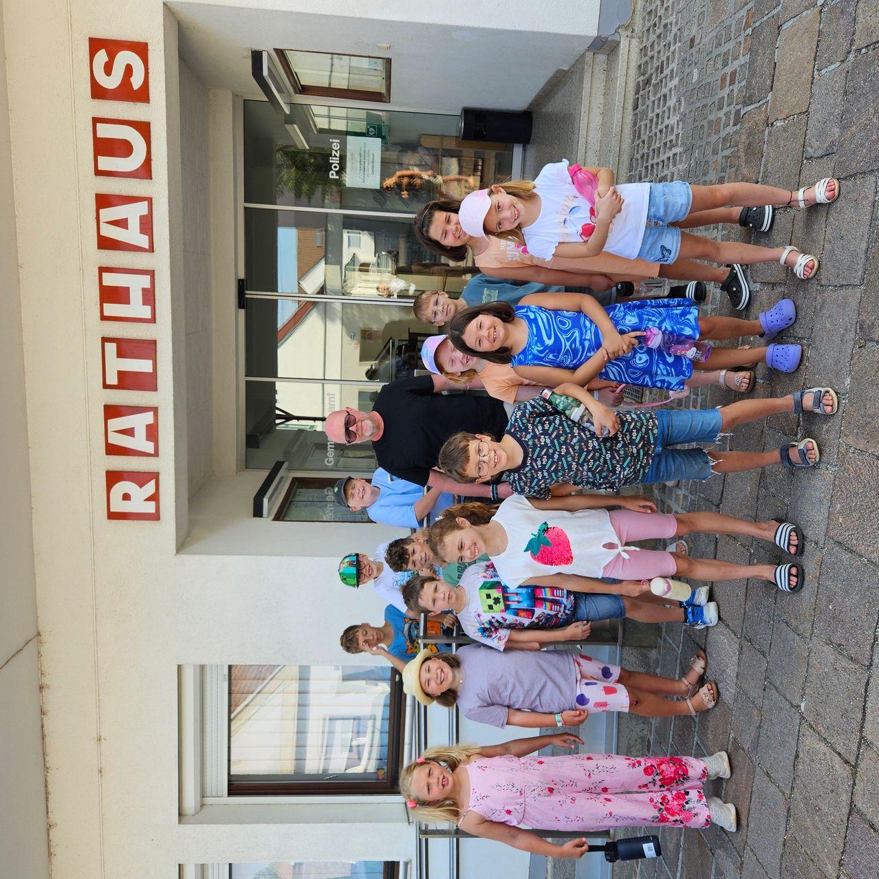 Eine Gruppe von Kindern und Erwachsenen steht vor einem Gebäude mit dem Wort Rathaus darauf. Sie lächeln und posieren für ein Foto.