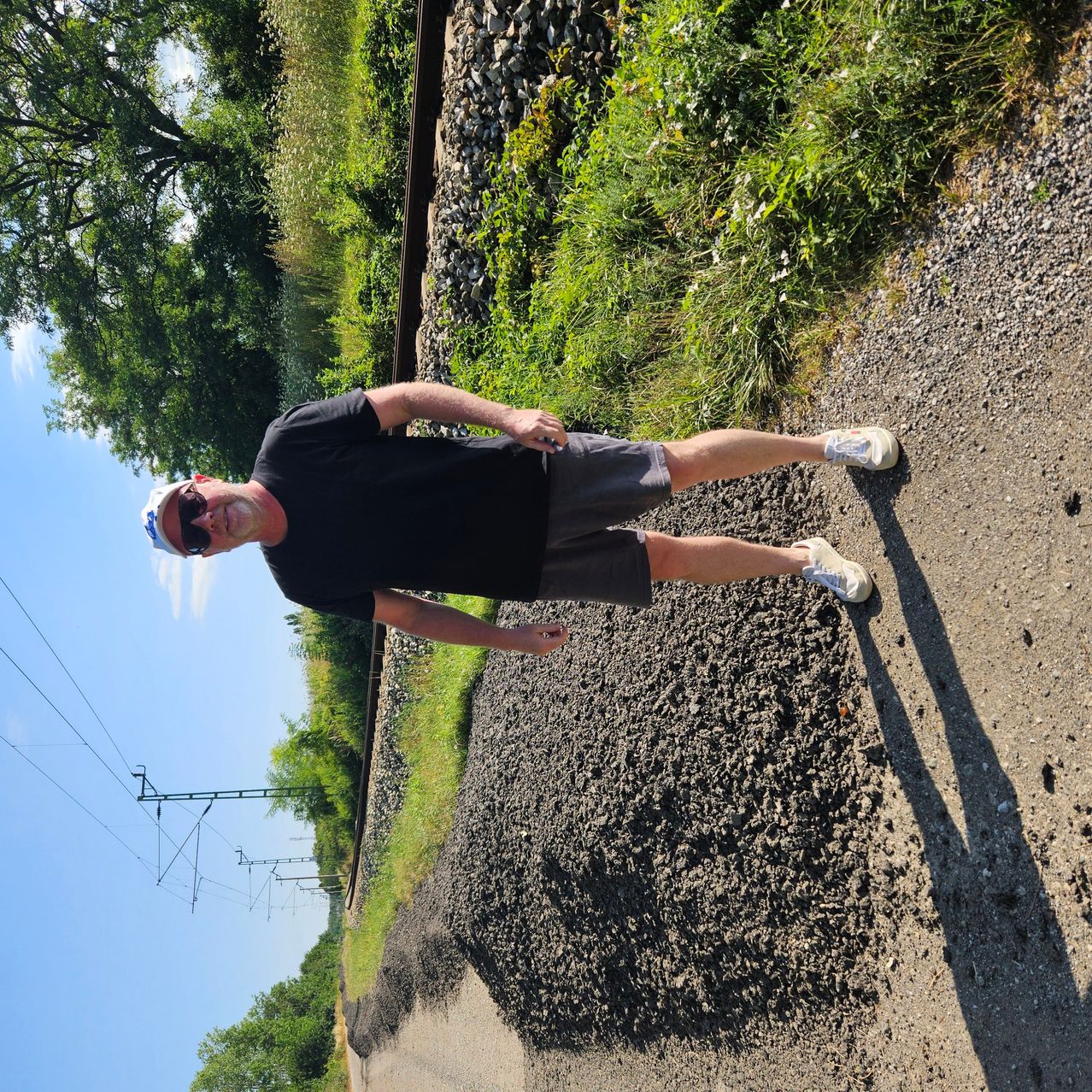 Ein Mann in einem schwarzen T-Shirt und Shorts posiert für ein Foto auf einem Schotterweg in der Nähe einer Bahnlinie, mit Grünflächen und Stromleitungen im Hintergrund.